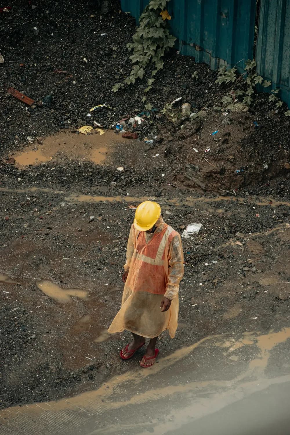 Un ouvrier en casque et vêtement de pluie sur un sol boueux couvert de flaques.
