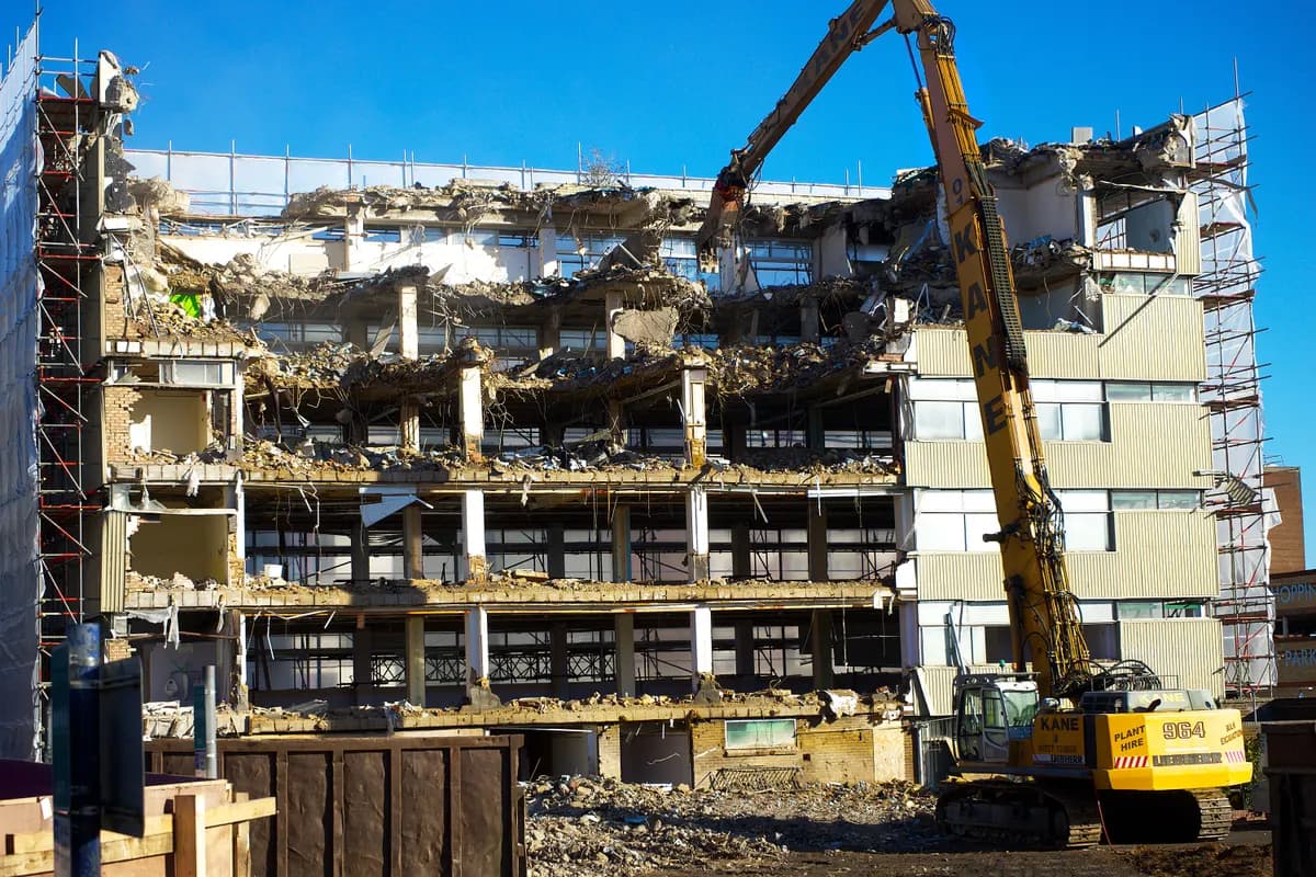 Une pelle de démolition intervient sur un bâtiment ouvert, entouré d’échafaudages.